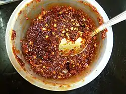 A red spread containing seeds in a white bowl, viewed top-down, with a metal spoon inside.