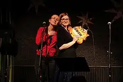 Photograph of the siblings almost back-to-back in front of a microphone stand, with Laser holding a cat-shaped electronic piano.