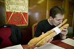 A jury member sits with his nose pressed up on a baguette, smelling it. Behind him is a banner of La Boulangerie de Paris.