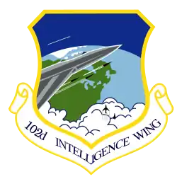 A standard shield, surrounded on the edge by a diminished yellow border. The upper part of the shield is blue, with one gray aircraft with a white trail pointing out from the left side. The plane is flying over the northeastern portion of the western hemisphere, which is colored in green and light blue. On the front of the gray aircraft with a white outline is a cockpit, with the jet firing three rockets. The three rockets trail white smoke and are aimed the same direction as the aircraft. Under the nose of the plane are two black aircraft coming out of clouds and rising up towards the jet. Attached to the shield is a white scroll edged with the same color border as the shield, surrounding the inscribed "102D INTELLIGENCE WING" in blue
