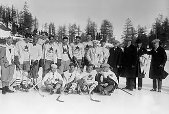 Black and white photo of fourteen hockey players in white sweaters with a maple leaf crest, and four men wearing dark suits and overcoats