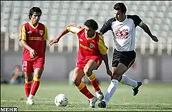3 football players competing for a ball, 2 having matching uniforms, "MEHR" is written in the bottom left corner