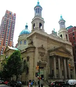 A stone building with a pediment and columns on the main entrance, two towers with green rounded tops and columns, and a dome at the rear lit by late afternoon sun from the right. There are traffic lights in front.