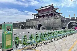 A line of green public bicycles next to a kiosk before a large historic gate