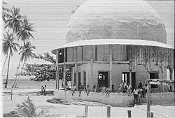 Black-and-white photograph, perhaps a photocopy, of a round dome on a beach. The dome is surrounded by a veranda with people sitting or standing looking at the viewer. Further there are 4 palm trees and another tree behind the dome. In the background, the sea.