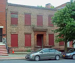 A two-story brick facade with red boards in the windows and brownstone around the entrance. Parked in front of a tree on the street is a dark gray late-2000s Honda Civic