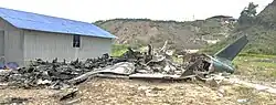 A burnt-out aircraft fuselage lying on dry ground next to a building