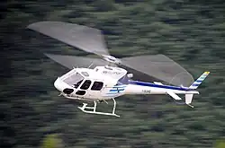 A white and blue helicopter in flight against a blurred forest background