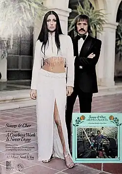 Cher stands in a white, fringed two-piece with long sleeves and a high-slit skirt, her long black hair parted in the middle. Behind her, Sonny Bono wears a black tuxedo with a bow tie, arms crossed. An inset in the bottom right shows their All I Ever Need Is You album cover.