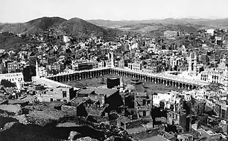 An old photograph showing a black, cubic structure enclosed by rectangular arcade surrounded by buildings and hills