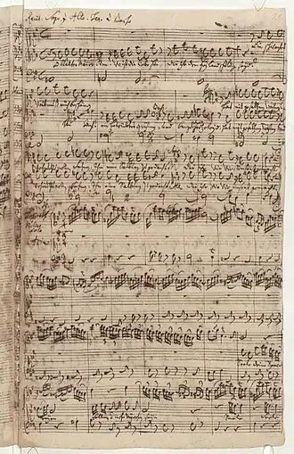A page of sheet music, an autograph of J.S. Bach, showing in 7 staffs the end of the first recitative, marked on top for four voices, and the beginning of an aria with an instrumental ritornello for a flute and basso continuo in the fourth staff, and the first phrases for the soprano voice in the sixths staff