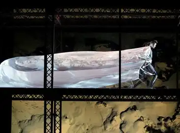 Model wearing extremely long cape walks through wind tunnel with cape billowing behind.