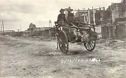 Two men in sombreros riding in a donkey-cart with a line of feet sticking out the back. They are riding down a dirt street away from the camera, with a line of buildings on the right. Dated 15 May 1911.
