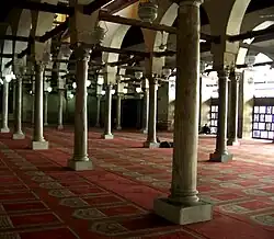 A large room filled with rows of cylindrical columns on top of square bases. The columns support arches which are pierced by square beams going the length of the room in both directions. Hanging from the beams are lamps, and the room's floor is covered with a red carpet with a repeated beige arched doorway shaped design on it. Exterior light enters from right of the room.