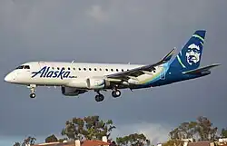 A plane painted with the words 'Alaska' in the front and a blue-green eskimo on the tail soars after take-off just under a cloudy sky
