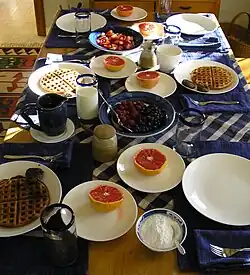 Table covered in mugs, glasses of milk, and plates of waffles, berries, powdered sugar, and grapefruit.