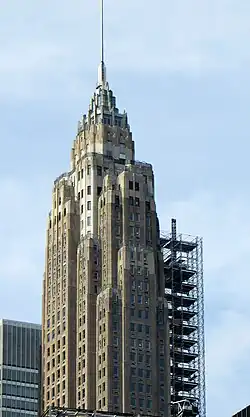 Upper section of the building, showing the small terraces on each setback. The top of the facade tapers to a metal spire. There is scaffolding outside the facade to the right.