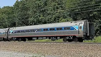 A corrugated silver tubular rail car with a blue stripe over the windows. At both ends, an angular light-blue shape covers the stripe.