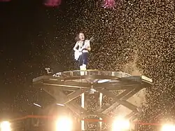 Angus, aged 55, stand atop a large, high pedestal. He is bare-chested and without his cap, he wears shorts while playing his guitar.