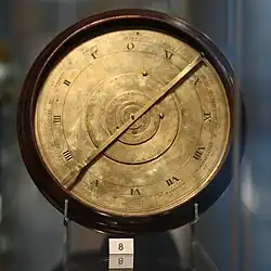 A brass, clock-like mechanical device in a museum display case, with a small card with the number 8 printed on it. The face of the device is split into several rings, with the Roman numerals I through XI (and 0) on one of these rings.