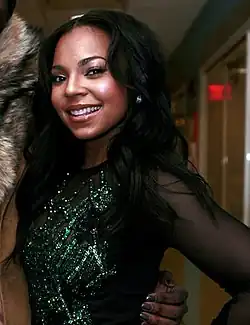 Ashanti in a somewhat dark room putting her hand on her left hip. She is looking directly at the camera while holding a smile.