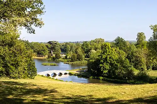 A colour photograph of the view over a lake from a hillside towards a building with tall chimneys, which is mostly hidden by trees
