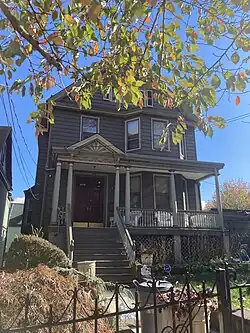 A three-story, gray-blue, Neo-Colonial style home with a gabled roofline, open porch, columns, and a pediment with a sunburst decoration. Picture includes shrubs and a trashcan on the front lawn, and the third story is partially obscured by leaves.