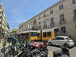An eletric bus and tram in Lisbon are stopped in traffic and blocking the street while other cars attempt to pass around them.