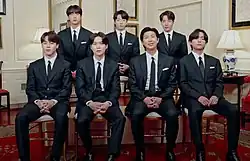 A group of seven South Korean men wearing suit and tie pose for the camera while sitting on their chairs