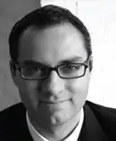 A black-and-white photograph of a man with dark hair and a cleft chin wearing glasses and looking at the viewer while smiling