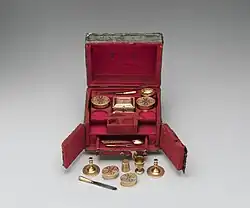 A small, elaborate box, featuring a hinged lid, two swing doors at the front and a small pull-out drawer; the interior is entirely red and features small items that seem to be part of a toilette set