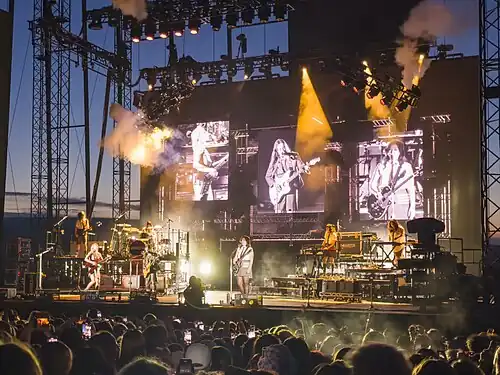 A brightly lit rectangular concert stage at sunset with puffs of smoke, television screens on the backdrop, and an audience watching
