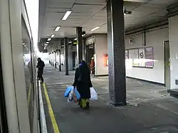 Three people walking on a platform, the one on the left boarding a train, the one in the middle carrying bags, and the one on the right walking through a door