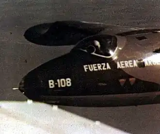 A close-up of a jet in flight, the pilot is wearing a white helmet. On the nose of the plane are the Spanish words "Fuerza Aerea Argentina" and the designation code "B-108".