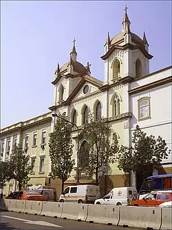 The image depicts a white church with two prominent towers. Surrounded by outdoor scenery, it is set against a sky with visible elements including trees and vehicles on the road.