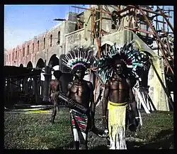 Two men with feather headdresses standing in front of an under-construction stone church