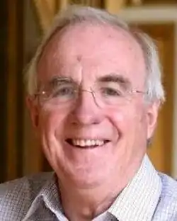 Colour Press shot of a smiling, clean-shaven, bespectacled man with short, receding, grey hair