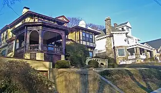 Two houses, one in a Tudor Revival style with a garage built into the ground beneath it and the other of wood with a stone foundation, seen from a street slopping downhill from them.