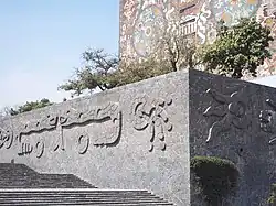 A large outdoor area with blackish-gray stone flooring of irregularly-cut medium-sized panels with mortar between them. Behind them in the distance are a grassy area with trees, buildings, and a blue sky with clouds