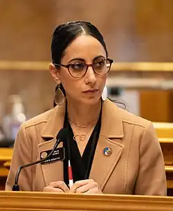 A woman in glasses and a tan jacket stands at a wooden podium, holding a pen. The background features ornate woodwork.