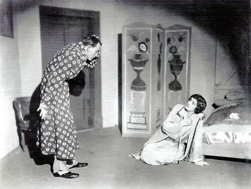 young man in a dressing gown looking down with a horrified expression at a middle-aged woman slumped to her knees on a bedroom floor looking back at him
