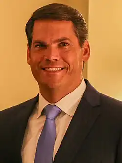 Portrait of Geoff Duncan in blue dress shirt with sleeves rolled up and red necktie, arms folded, in front of a U.S. flag.