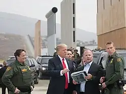 Trump is speaking with U.S. Border Patrol agents. Behind him are black SUVs, four short border wall prototype designs, and the current border wall in the background.