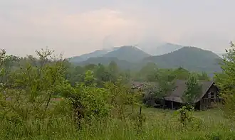 A green landscape with mountains in the back and a house towards the right.