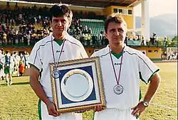 Two men in white football jerseys holdin a plaque.