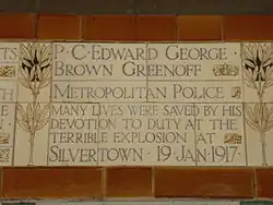 A tablet formed of five tiles of varying sizes, bordered by yellow wheatsheaves in an art nouveau style. The tablet reads "P.C. Edward George Brown Greenoff, Metropolitan Police. Many lives were saved by his devotion to duty in the terrible explosion at Silvertown 19 Jan 1917".