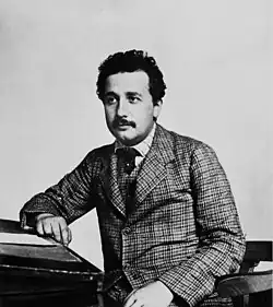 Head and shoulders shot of a young, mustached man with dark, curly hair wearing a plaid suit and vest, striped shirt, and a dark tie.