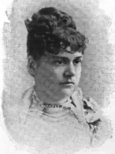 Portrait of a white woman with dark curly hair in an updo. She is wearing a light-colored garment, with beads at her throat.