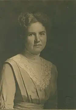 A white woman with greying hair, wearing a dress with a pleated bodice and a lace bib detail