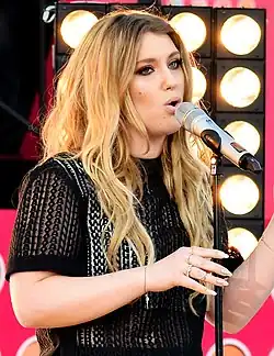 Young woman with long light coloured wavy hair wearing black clothing singing at a microphone